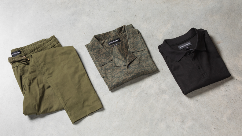 A pair of green pants, a camouflage camp shirt, and a black polo on a tabletop.