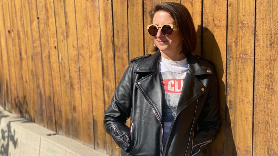 A woman stands against a wooden fence wearing a black leather biker jacket.