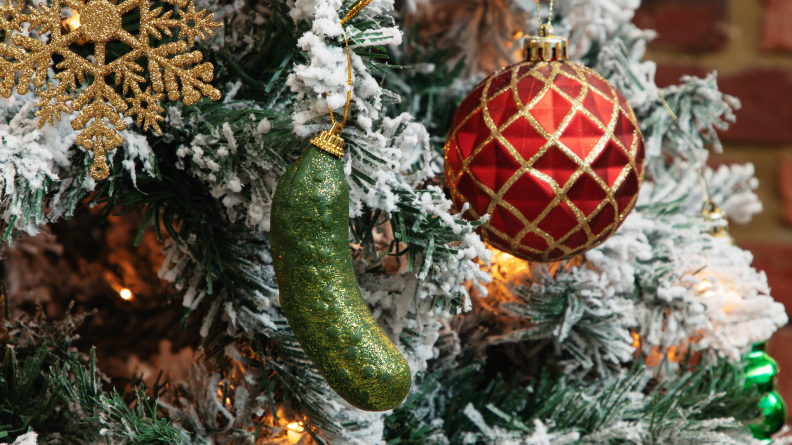 A Christmas pickle ornament on a flocked Christmas tree with a red ornament in the background.