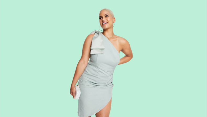 A woman wears a sage green, figure-hugging midi-length dress with a bow on the shoulder and a slit along the thigh.