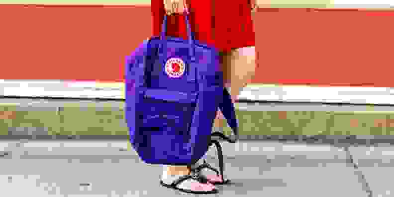 A person holds a purple backpack while walking on a sidewalk.