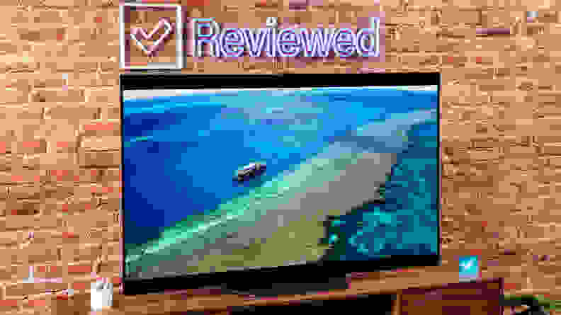 The LG B2 on a wooden table in front of a brick wall with a neon Reviewed sign displaying a beach.