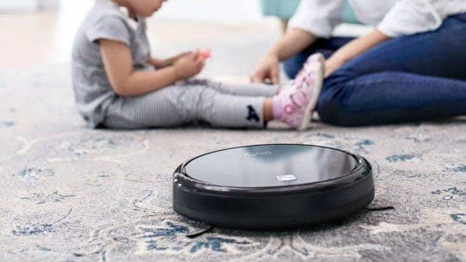 A Eufy 11S robot vacuum cleaning a carpet with a woman and child sitting in the background.