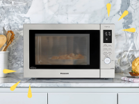 A Panasonic microwave sits on a marble kitchen counter surrounded by cooking utensils and fresh vegetables.