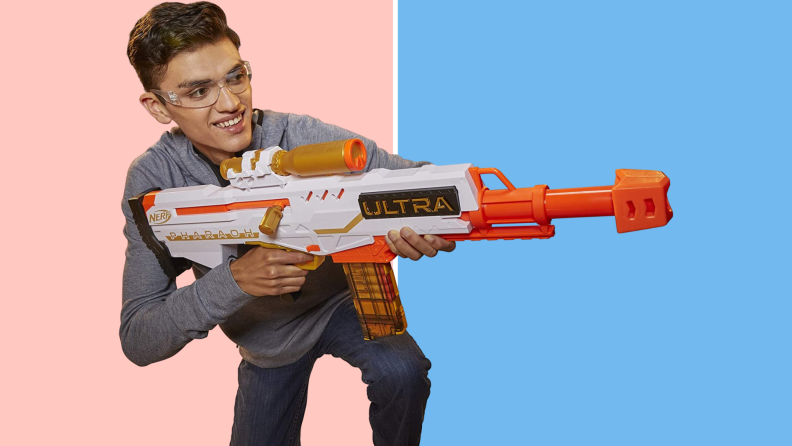 A teen in safety goggles holds the Nerf Ultra Pharaoh Blaster against a pink and blue background.