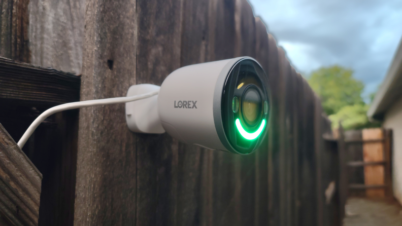 Lorex 4K Spotlight mounted on wooden fence outdoors with green LED light.