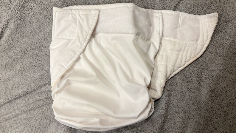White diaper with hook-and-loop closure, one flap open but otherwise closed against a gray fabric surface,