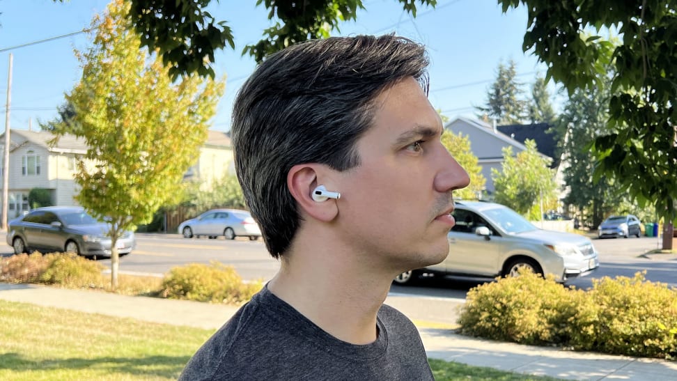 A man wears the AirPods Pro 2 earbuds while outside in a neighborhood park on a sunny day with cars going by in the background.