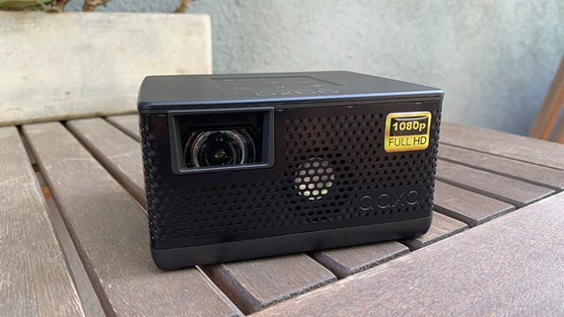 The AAXA P400 portable projector sitting on a wooden table outdoors in front of a stucco wall.