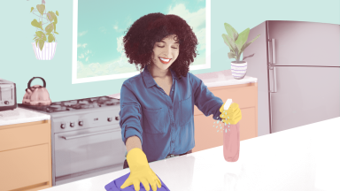 Person with curly hair smiling while cleaning countertop surface with rubber gloves and cleaning solution in kitchen.