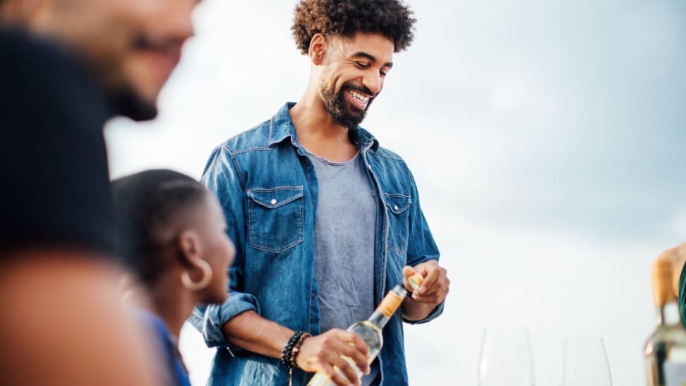 A smiling man opens a bottle of white wine in front of his friends