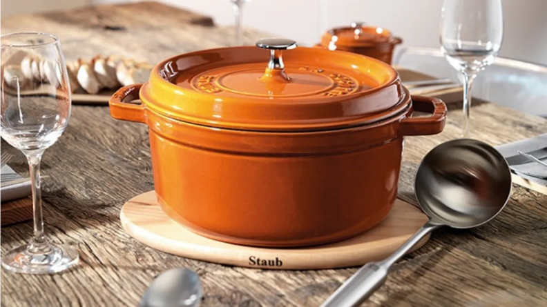 An orange Staub Dutch Oven on a table next to wine glasses, a steel spoon, and plates.