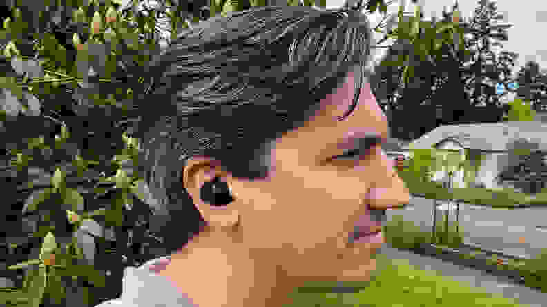 A black model of Sennheiser's Momentum TW 3 earbuds sit in a man's ear in front of green bushes and a green lawn.