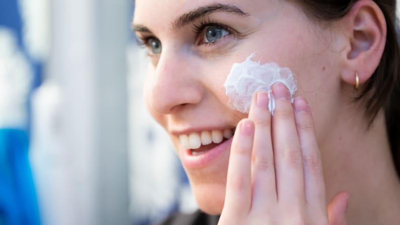 A person smiling and applying white face moisturizer to her cheek.