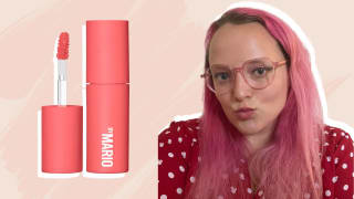 On the left: A coral-colored doe-foot applicator and lipstick tube. On the right: A person with pink hair wearing a red top and glasses purses their lips, which has a pink color on it.