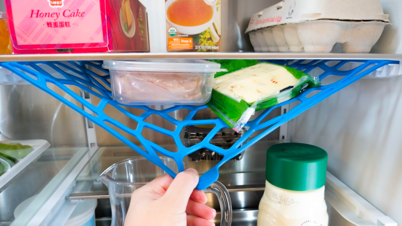 A close-up of a blue silicon web that's stretched out underneath a fridge shelf. A hand is reaching into frame to pull down its edge, showcasing how some deli meats and sliced cheese fit in the net.