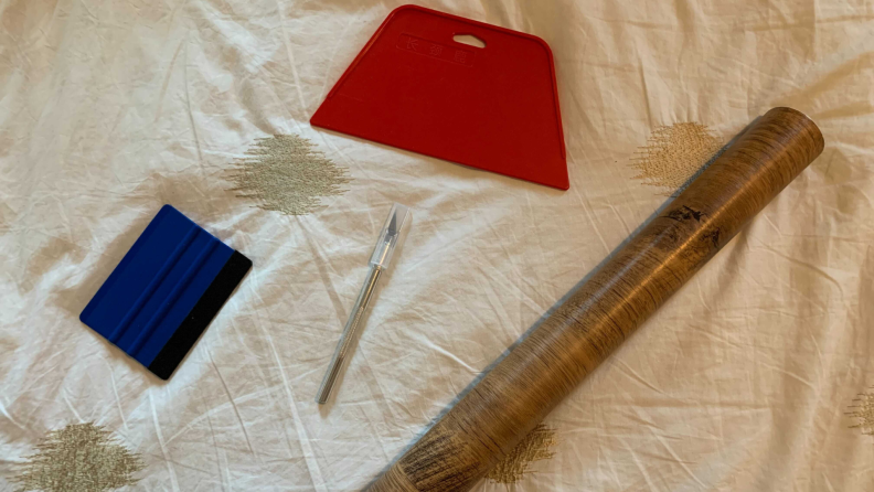 One large red smoothing tool, one small blue smoothing tool with a soft felt edge, one craft knife, and a roll of wood-textured contact paper