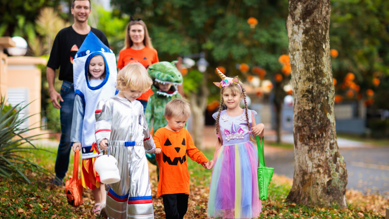 Children in Halloween costumes walk in a group together to trick-or-treat as the parents supervise them outdoors.