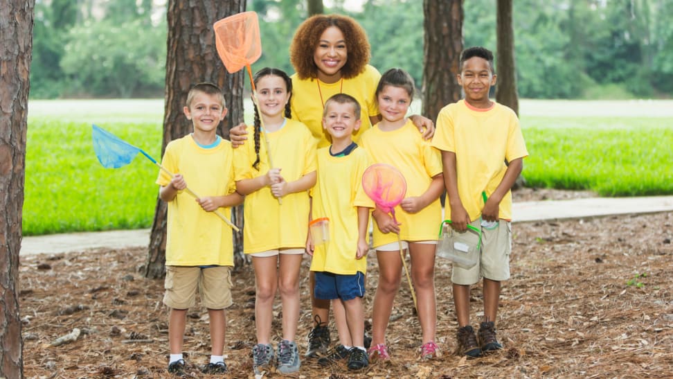 A group of five young children are joined by their camp counselor outside.