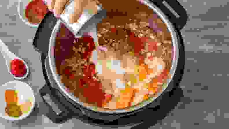 A hand pours ingredients into a pressure cooker that is already partially filled with other ingredients.