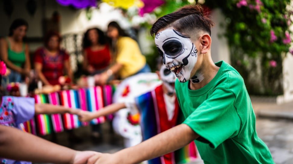 Small children with skull painted faces playing together while holding hands in a circle at a Dia de Los Muertos event outdoors.