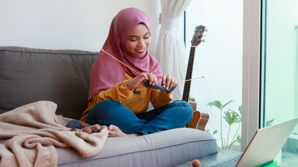 Woman on couch knitting with yarn while looking at laptop