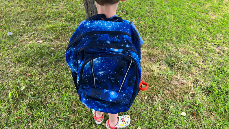 Small child wearing Lands' End ClassMate XL backpack in Blue Galaxy Space color pattern outdoors on grass.