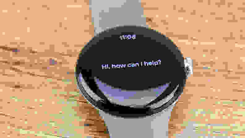 A Google Pixel Watch sits on a counter top displaying the message 'How can I help?'