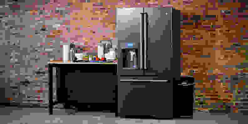 A modern fridge stands against a brick wall, flanked by a table and trashcan.