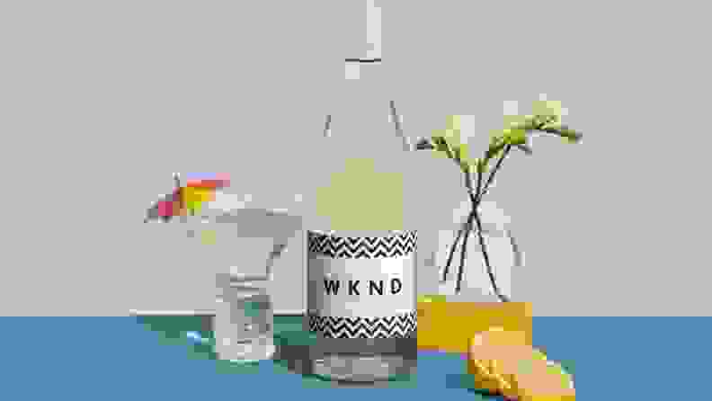 An artistic image featuring a bottle of white wine, a wine glass filled with the white wine and topped with a decorative umbrella, plus some lemon sliced and a small vase with flowers on the side.