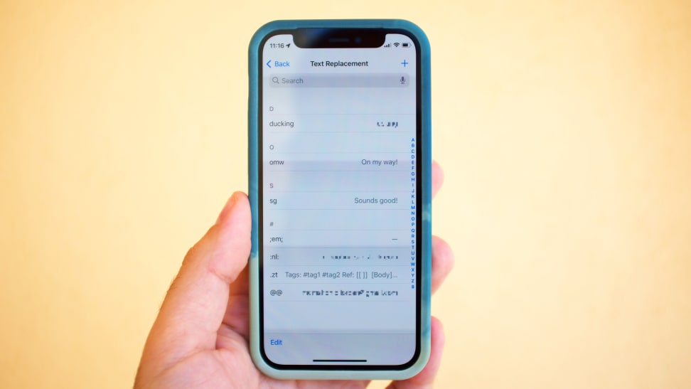 A hand holding an iPhone 12 Mini, inside a green case, with iOS's Text Replacement feature on-screen. There are several shortcuts shown on-screen, including two @ symbols, which expands into an email address.