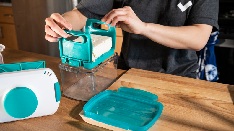 A person pulling a block of tofu out of the Noya Tofu Press, using the handles on the removable drip tray.