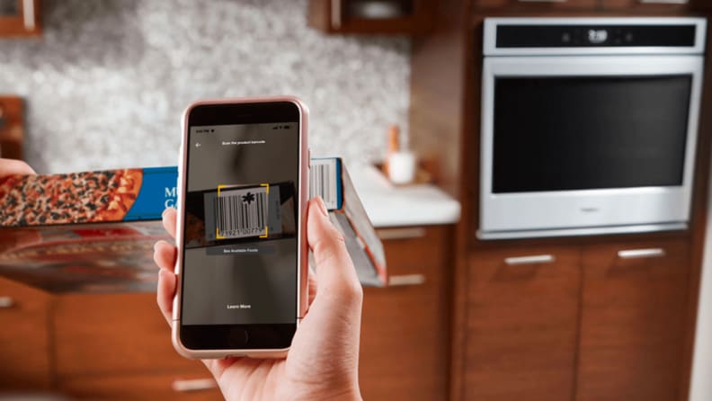 A hand holds a smart phone up to scan the QR code of a frozen food box. In the background is a Whirlpool smart oven, which is presumably receiving cooking information from the app and preheating to the appropriate temperature.