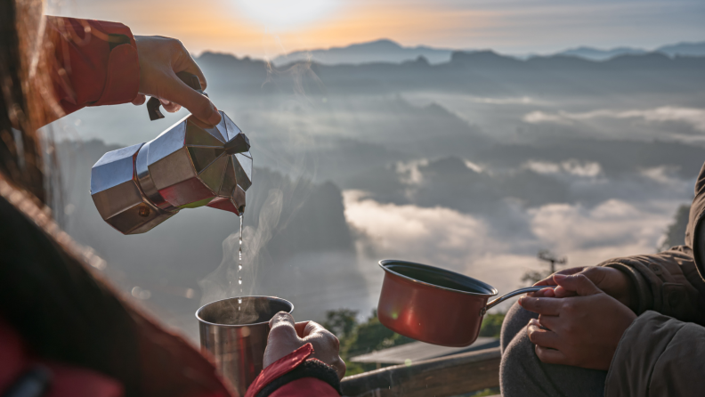 A person is holding a moka pot and pouring liquid to a mug with another person with a small pot next to her.