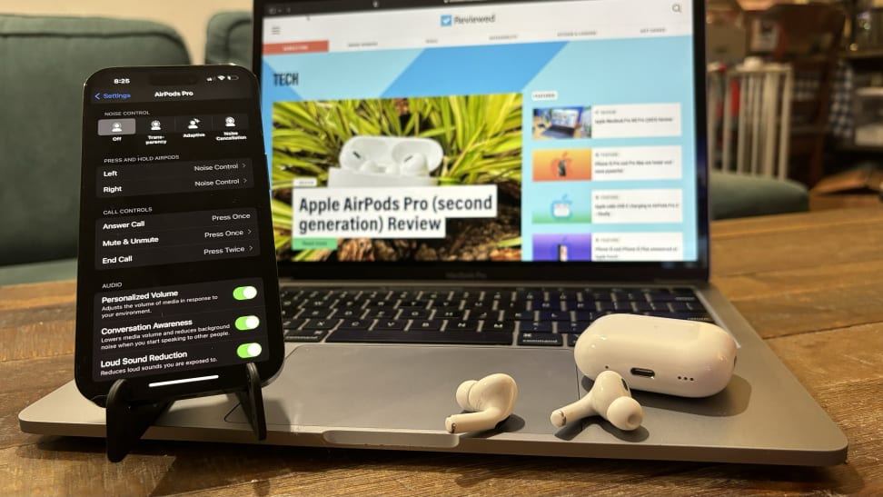 An iPhone 14 Pro displaying the Apple AirPods Pro 2 menu, sitting on a Macbook Pro with the Apple AirPods Pro 2 earbuds and case next to it, all sitting on a wooden coffee table.