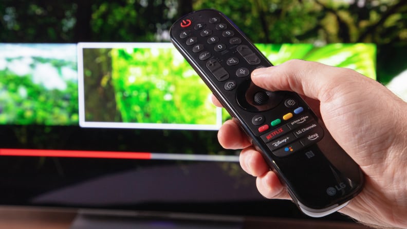 A close up of a hand holding the LG Magic Remote with a TV in the background.