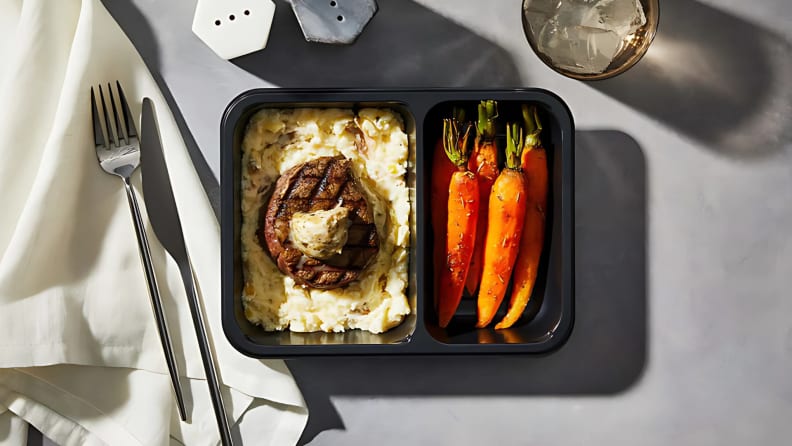 A prepared Factor meals entrée featuring grilled steak, mashed potatoes, and roasted carrots in a black tray.