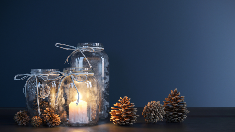 Three frosted mason jars with tea lights inside and pinecones on the table next to the jars