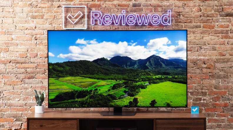 Samsung QN90D Mini-LED TV with color rich scenery on screen atop a wooden TV stand below Reviewed neon sign in front of brick wall indoors.
