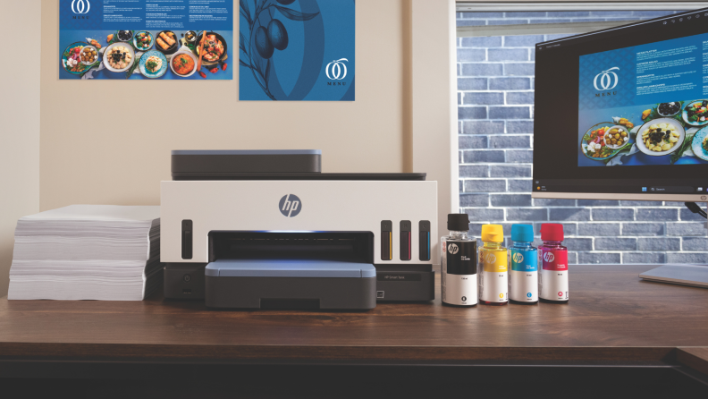 HP Smart Tank printer on a desk with ink bottles and a computer monitor displaying a menu design.