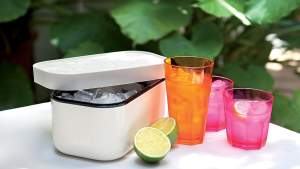 A white Lekue ice box filled with ice cubes sits open on a table beside three drink glasses filled with ice. Two halves of a lime are in the foreground.