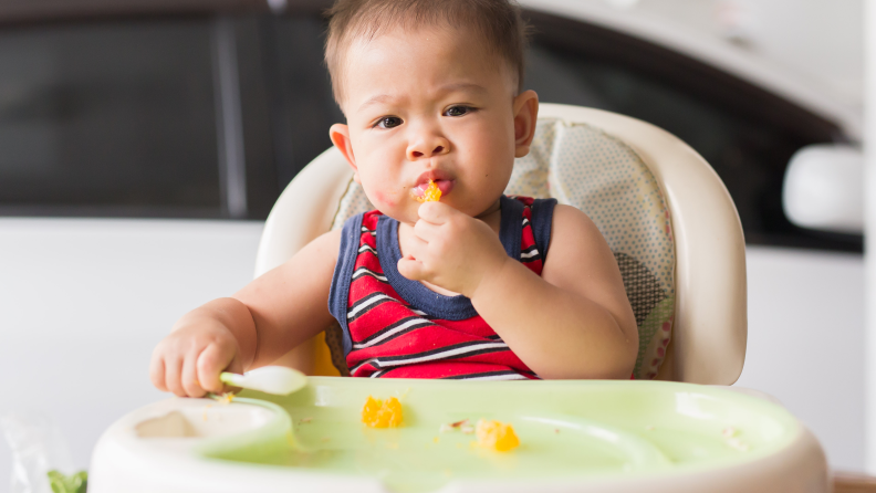 A baby boy in a high chair eating an orange and holding a spoon