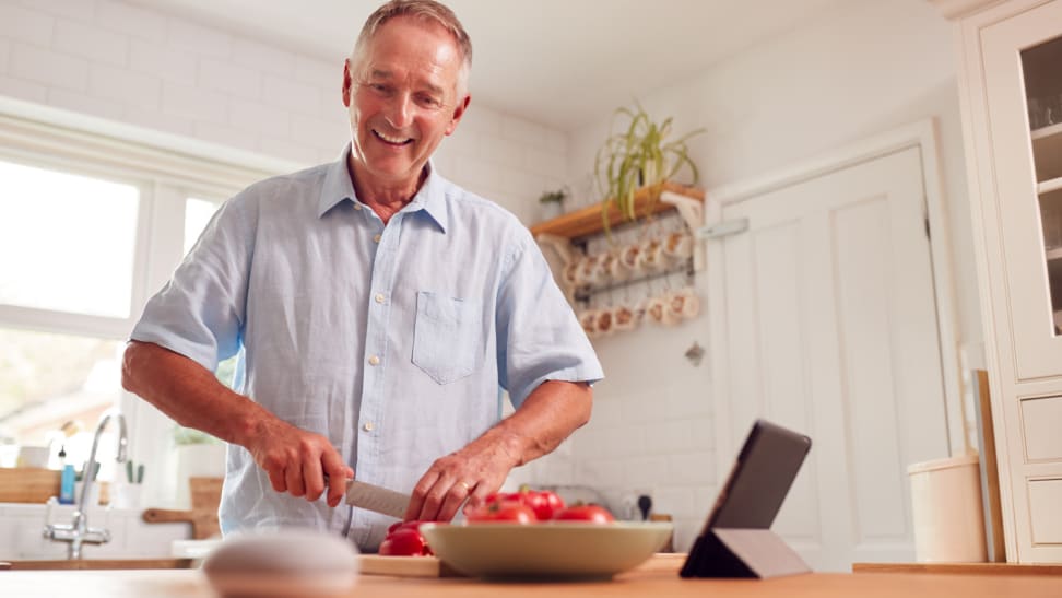 A person chops tomatoes in a kitchen and listens to a smart speaker.