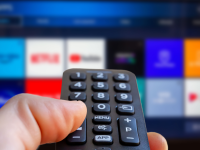 A man is holding a remote control of a smart TV in his hand. In the background you can see the television screen with streaming entertainment apps for video on demand.