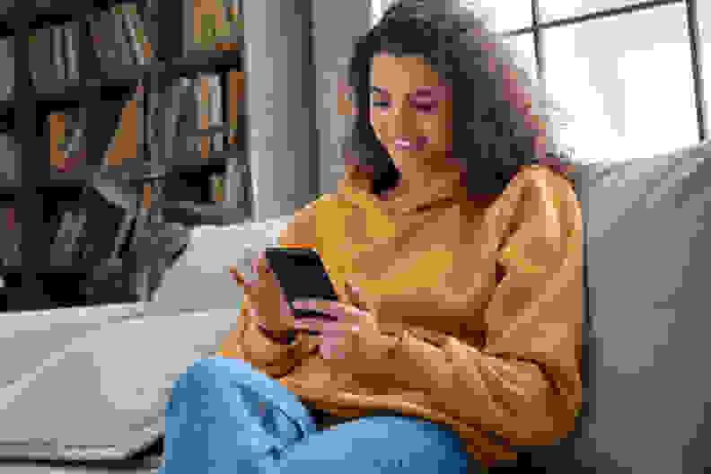 快乐的千禧西班牙少女在家拿着智能手机查看社交媒体。微笑的年轻拉丁女子使用手机app玩游戏，网上购物，订购送货在沙发上放松。