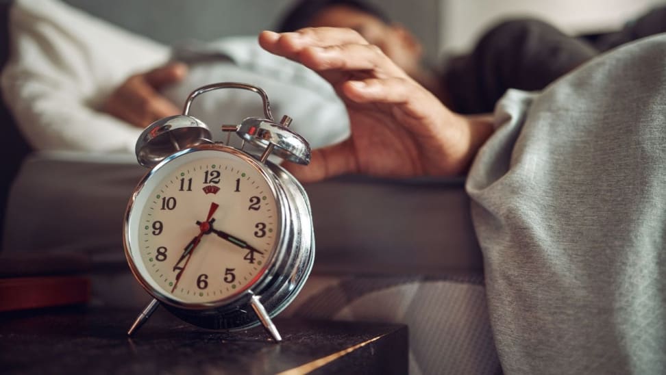 Person sleeping in bed next to silver alarm clock on night stand.