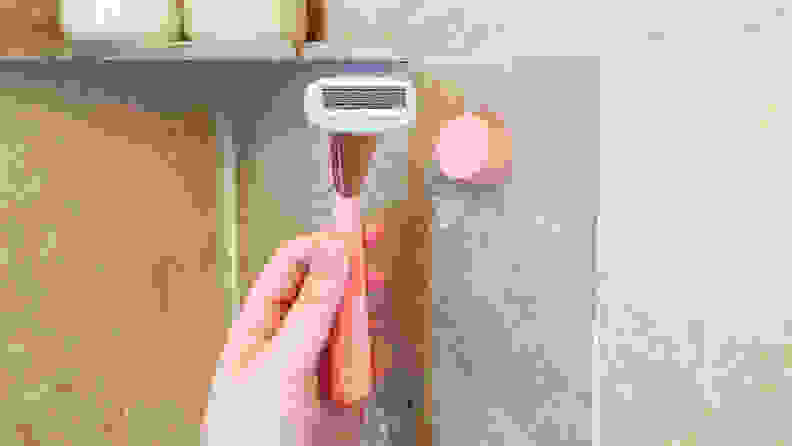 A hand holding a pink Athena Club Razor in front of its wall mount, which is stuck to the shower wall.