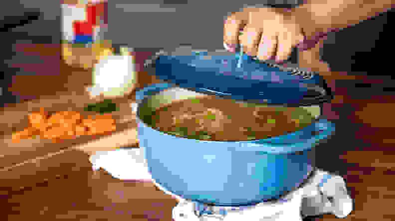 A person lifts the lid of a blue Dutch oven to reveal chicken soup inside.