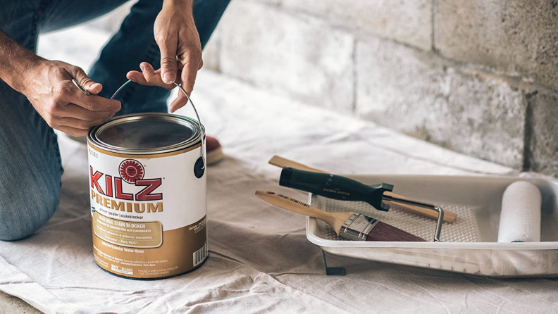 A person opens a can of white primer that's sitting next to painting tools.