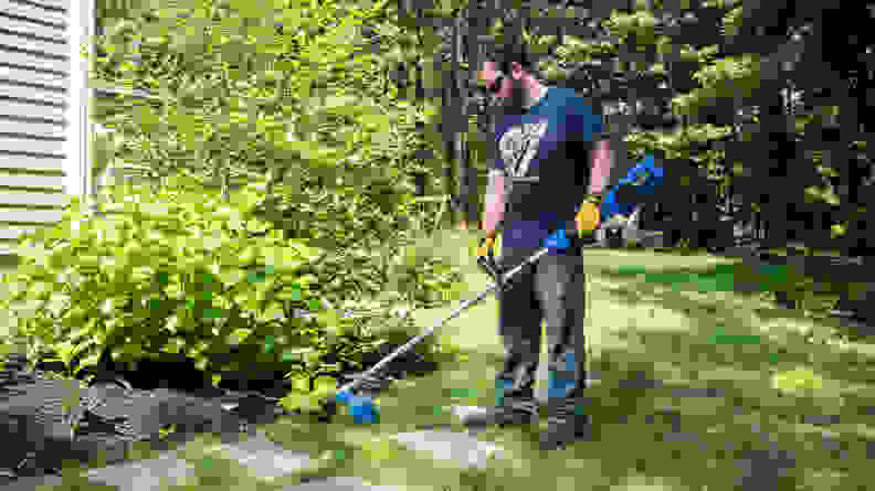 A man uses a weed wacker to cut grass around the edge of a garden